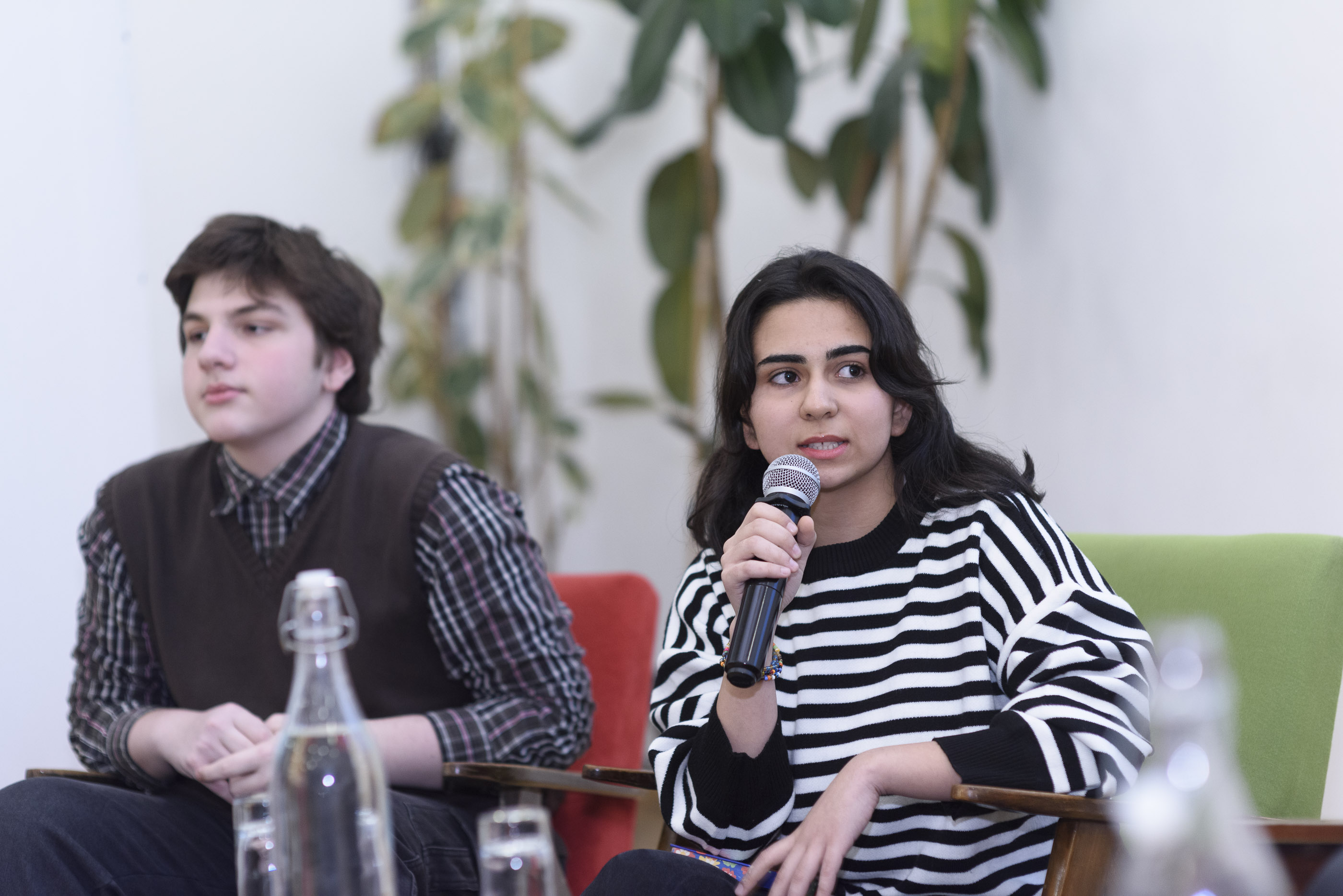 A girl and a boy are sitting. The girl is holding a mic, speaking