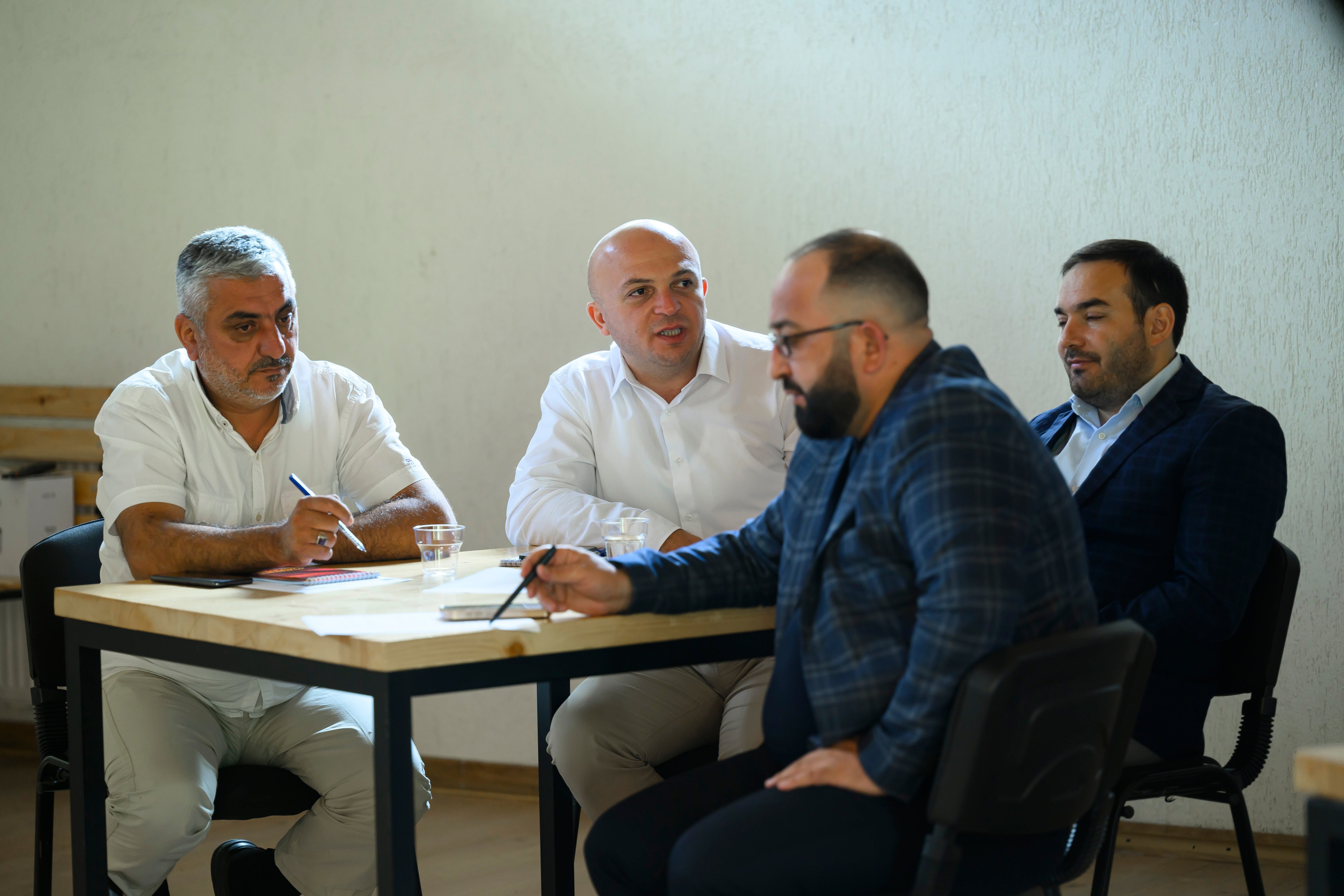 Four men sitting at a table. Two of them are wearing while, while the others are wearing dark suits