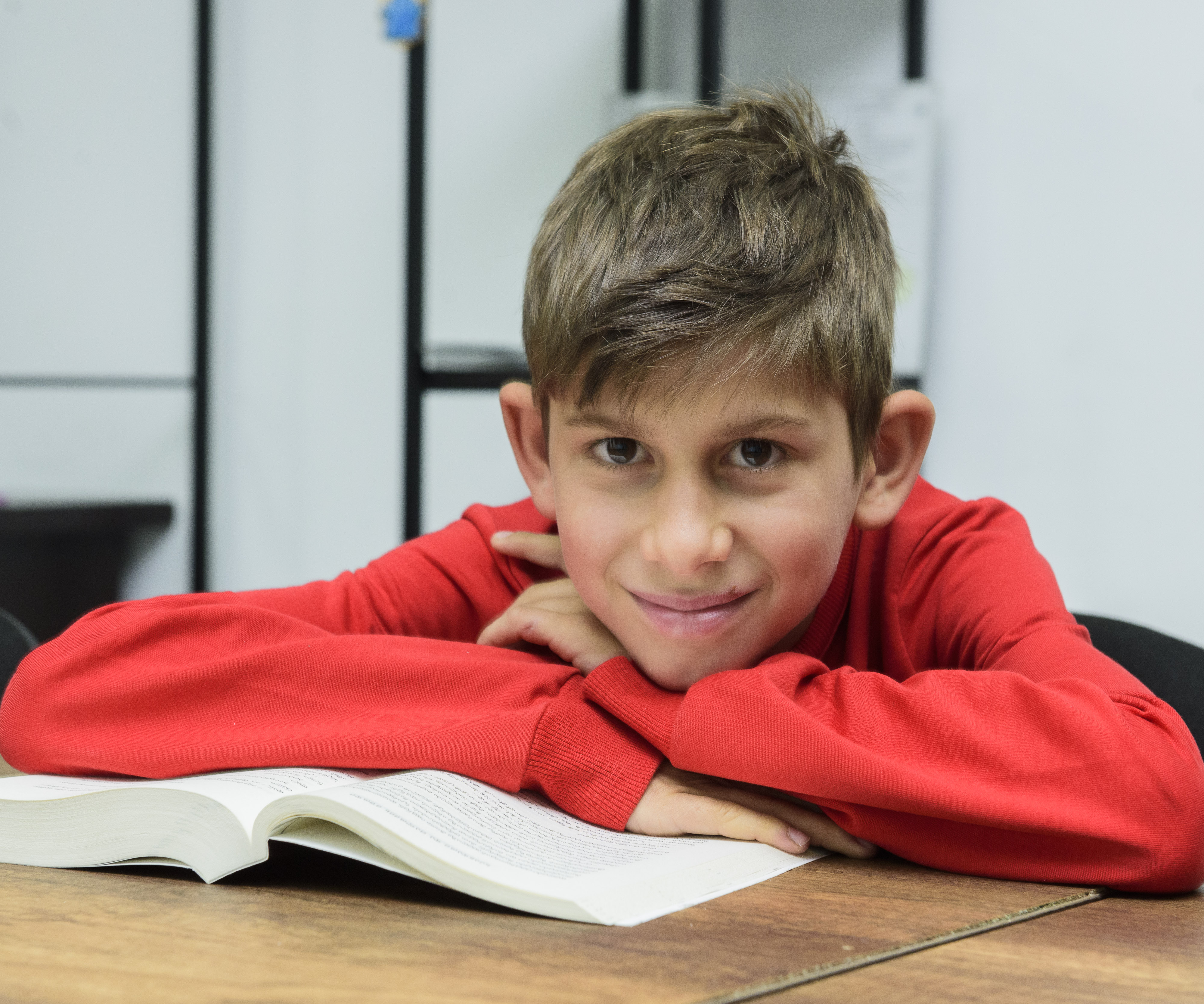 An adolescent boy wearing a read long-sleeve shirt is leaning on a book smiling