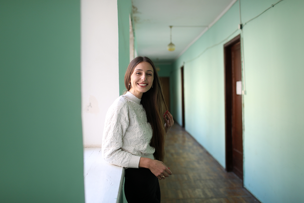 Ana Datiashvili standing at a window of a building wearing a white long-sleeve shirt and black trousers