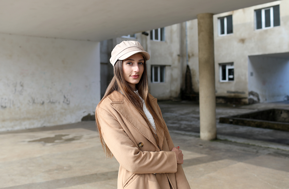 Ana Datiashvili wearing a white cap and light brown coat is standing outside a building