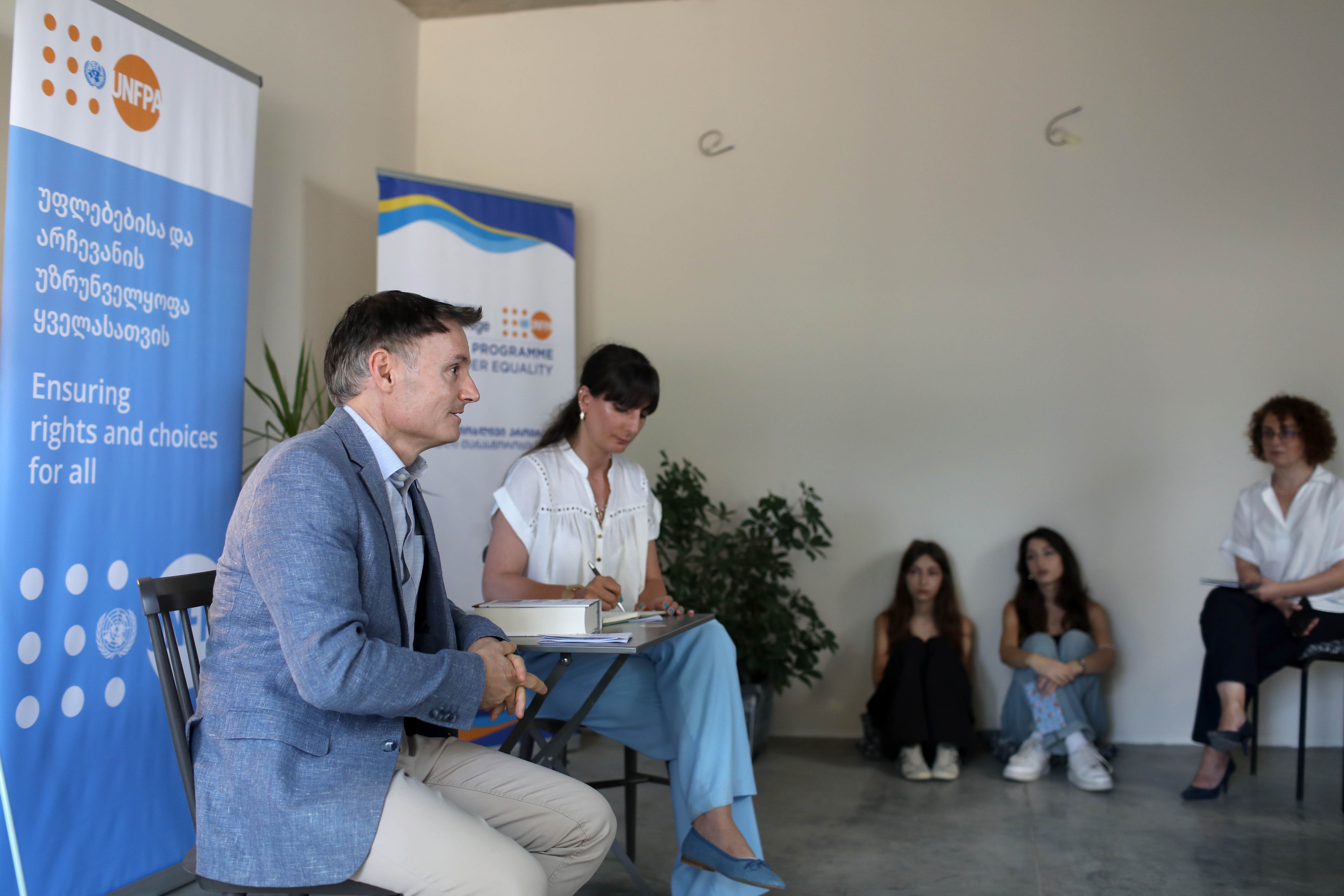 Two people sitting at a panel, while two adolescent girls are sitting at a wall, listening