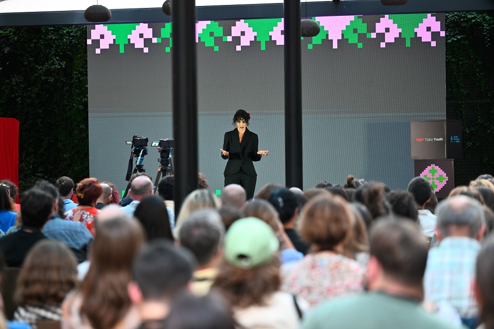 Lizi Katamadez, wearing black and standing on a stage is addressing the audience in front of her