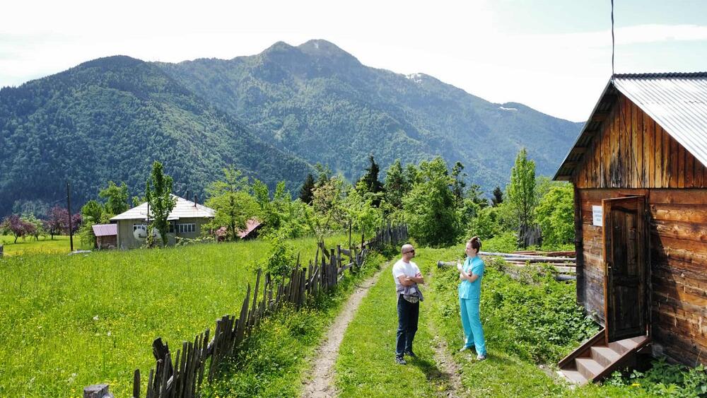 A scene of a village in upper Adjara. A young woman and a man are talking in front of an old wooden house
