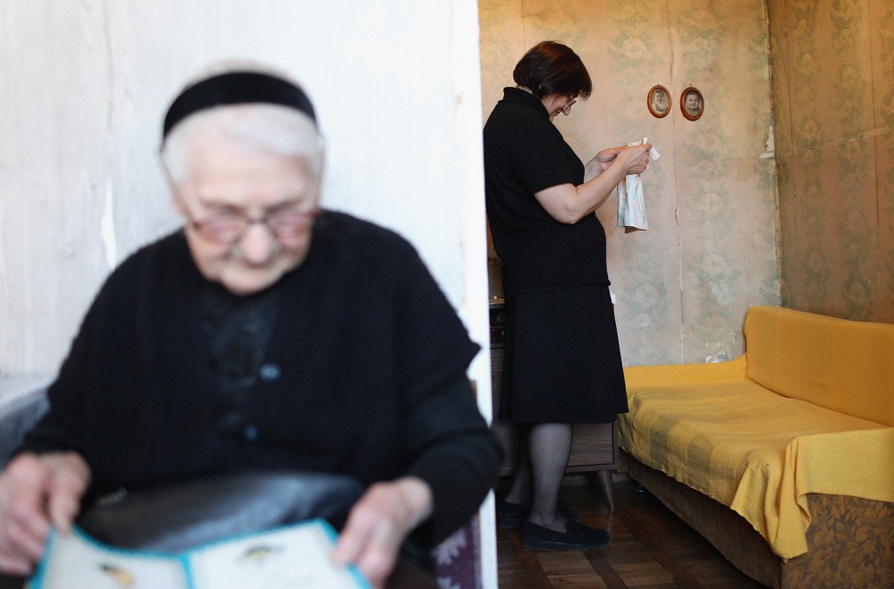 An elder woman in black is looking into something. There is another woman behind her, standing