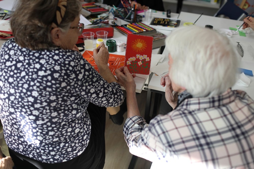 Two women sitting from their back. One of them is showing to another the flower that she has painted