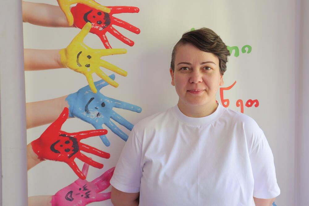 A woman with short hair, wearing white T-shit is standing in a room in front of a wall with colorful hands painted on the wall