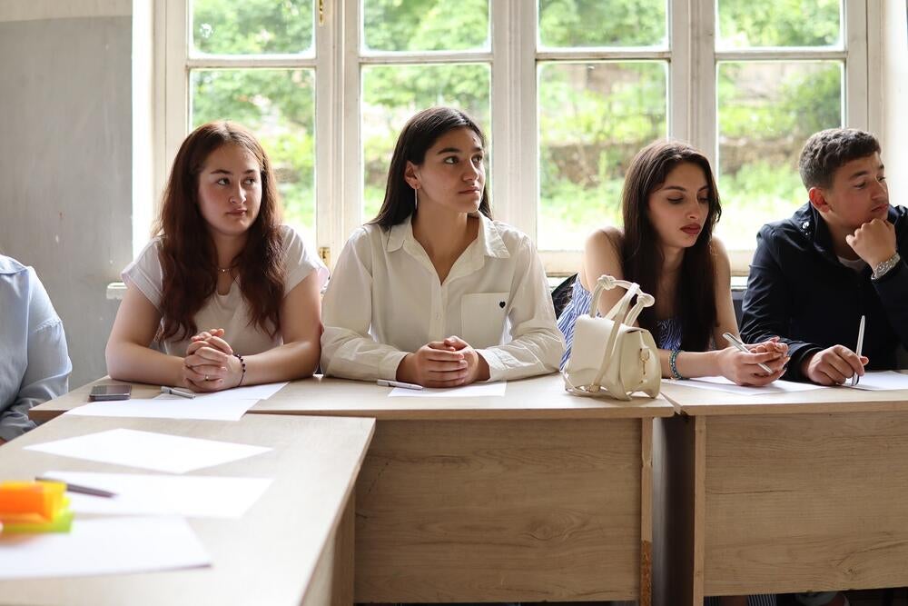 Three adolescent girls and a boy are sitting in a classroom in front of a window