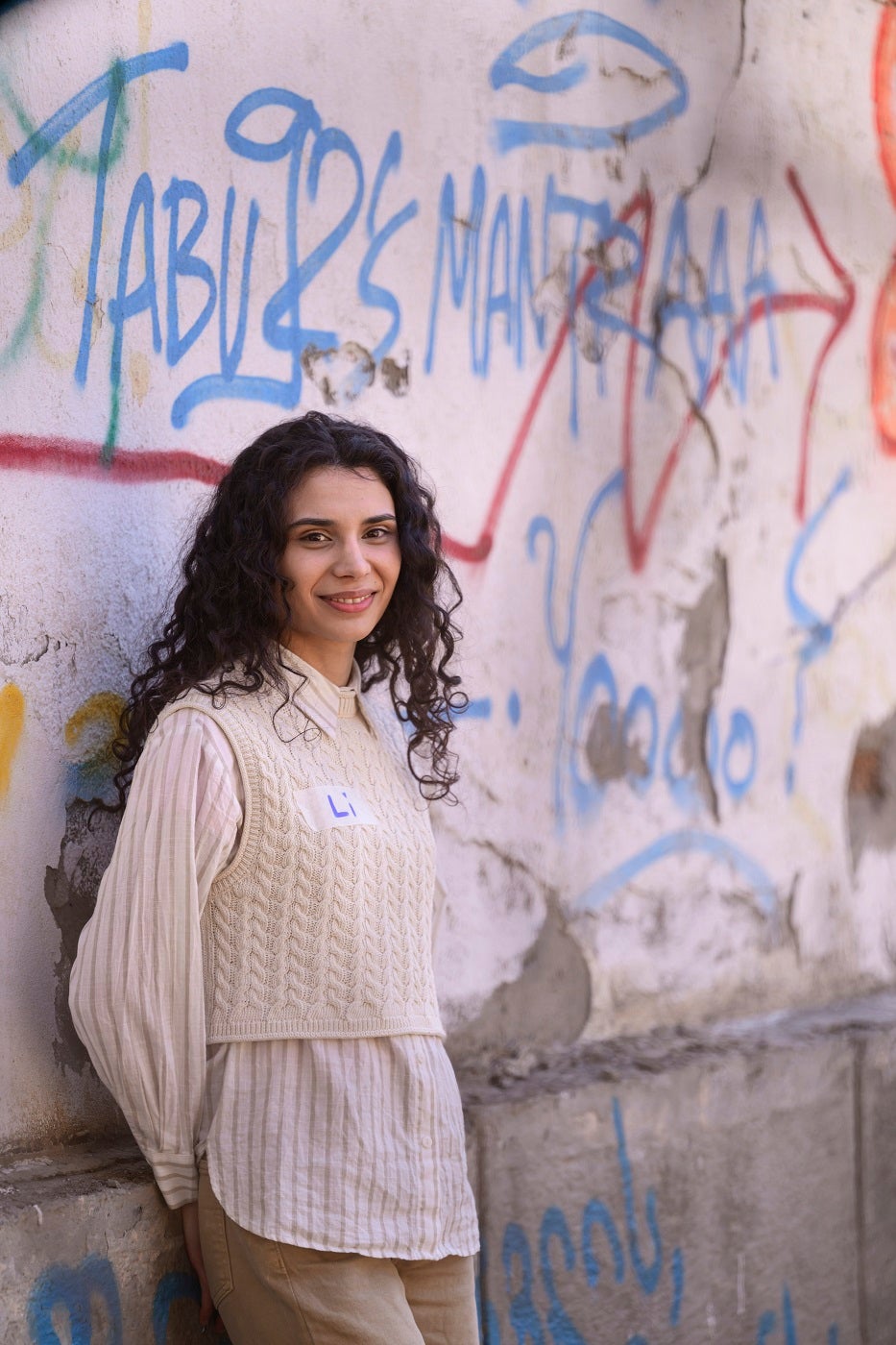 A young woman with curly hair and white shirt is standing at a wall smiling