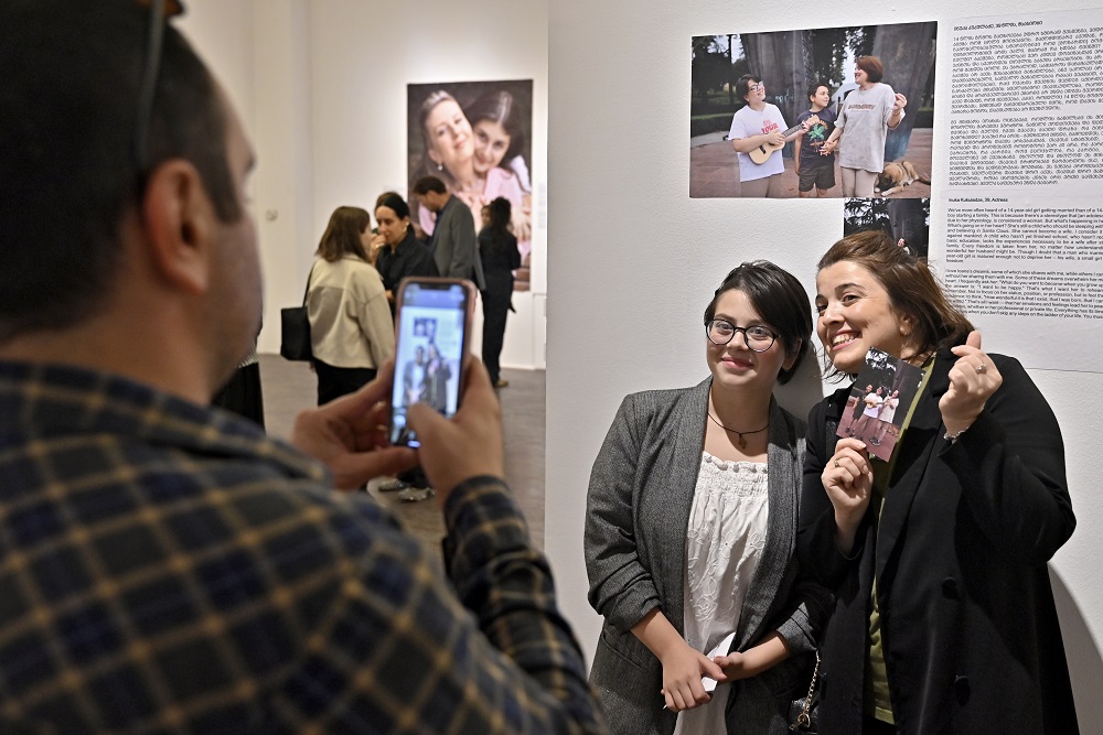 A woman and her daughter standing at a wall, holding their photo, while a man is photographing them