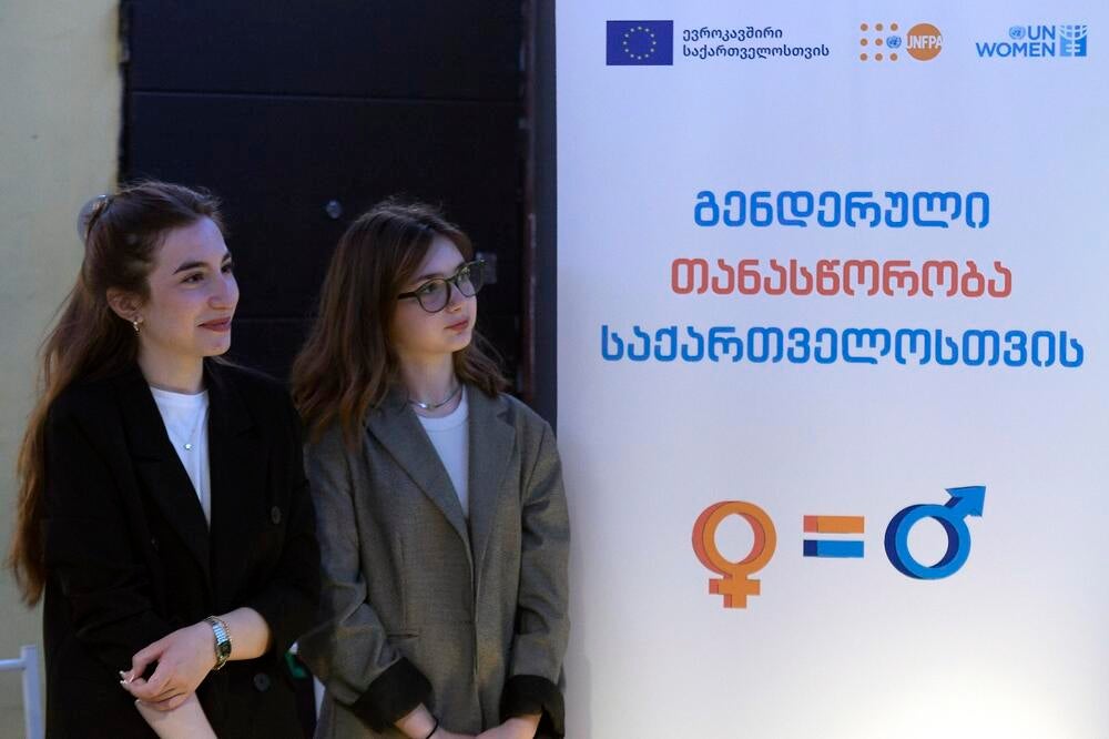 Two young women standing at a banner with the European Union, UNFPA and UN Women logos and a Georgian title of the programme: Gender Equality for Georgia