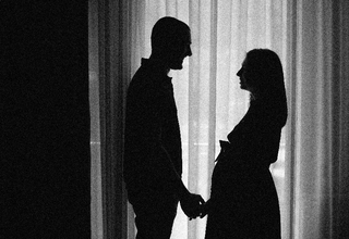 A black and white photo of a couple, expecting a baby standing near the curtains holding hands
