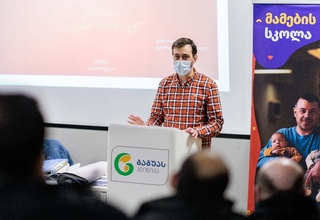 A man wearing a mask is standing in front of a large screen which reads: MenCare campaign. There is a rollup banner of Fathers' Sshool on the left side of the photo