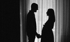 A black and white photo of a couple, expecting a baby standing near the curtains holding hands