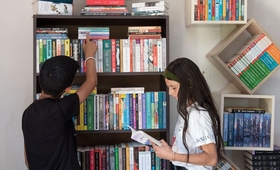 A boy and a girl are standing at a bookshelf. The boy is looking for a book, while a girl is holding one