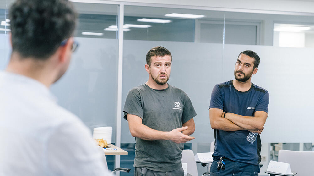 Three men standing and talking. One of them wearing a white shirt is seen from the back, while other two are standing in front