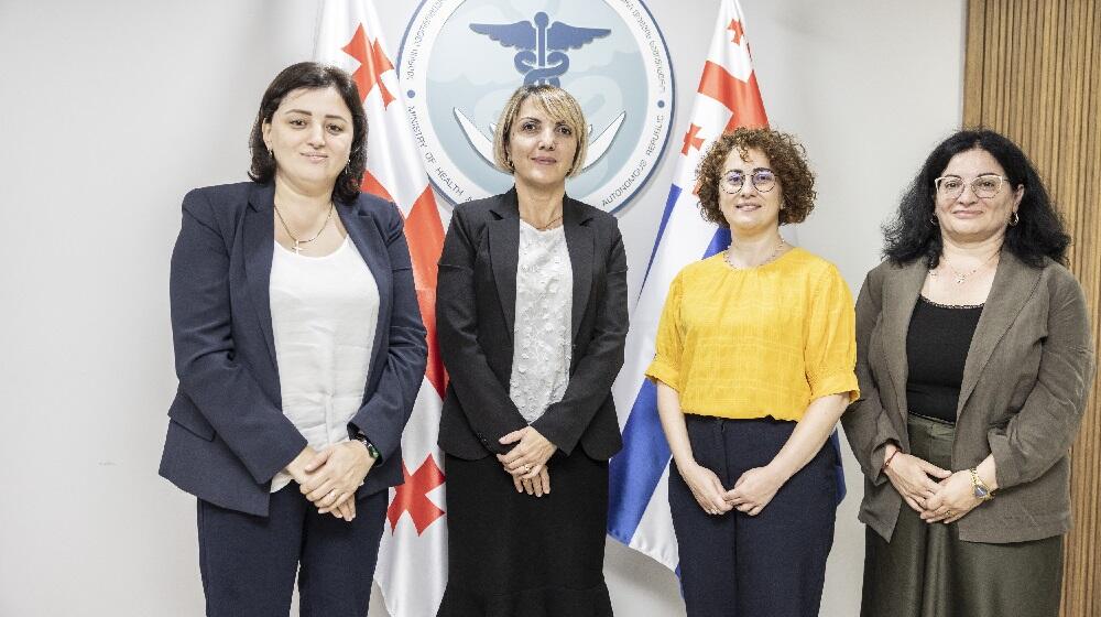 Four women standing together in front of a large logo of the health ministry and two flags of Georgia