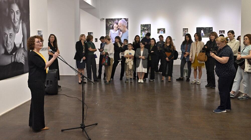 A bunch of people standing in a circle at a photo wxhibition listening to the Head of UNFPA Georgia Country Office, Mariam Bandzeladze, speaking in a microphone