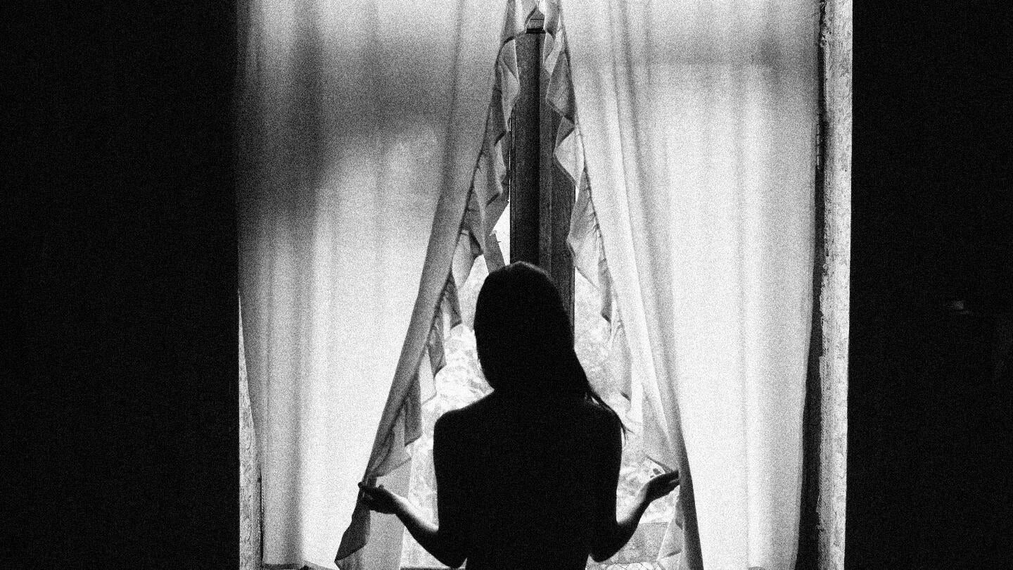 A black and white photo of a woman standing at a window hoilding curtains open