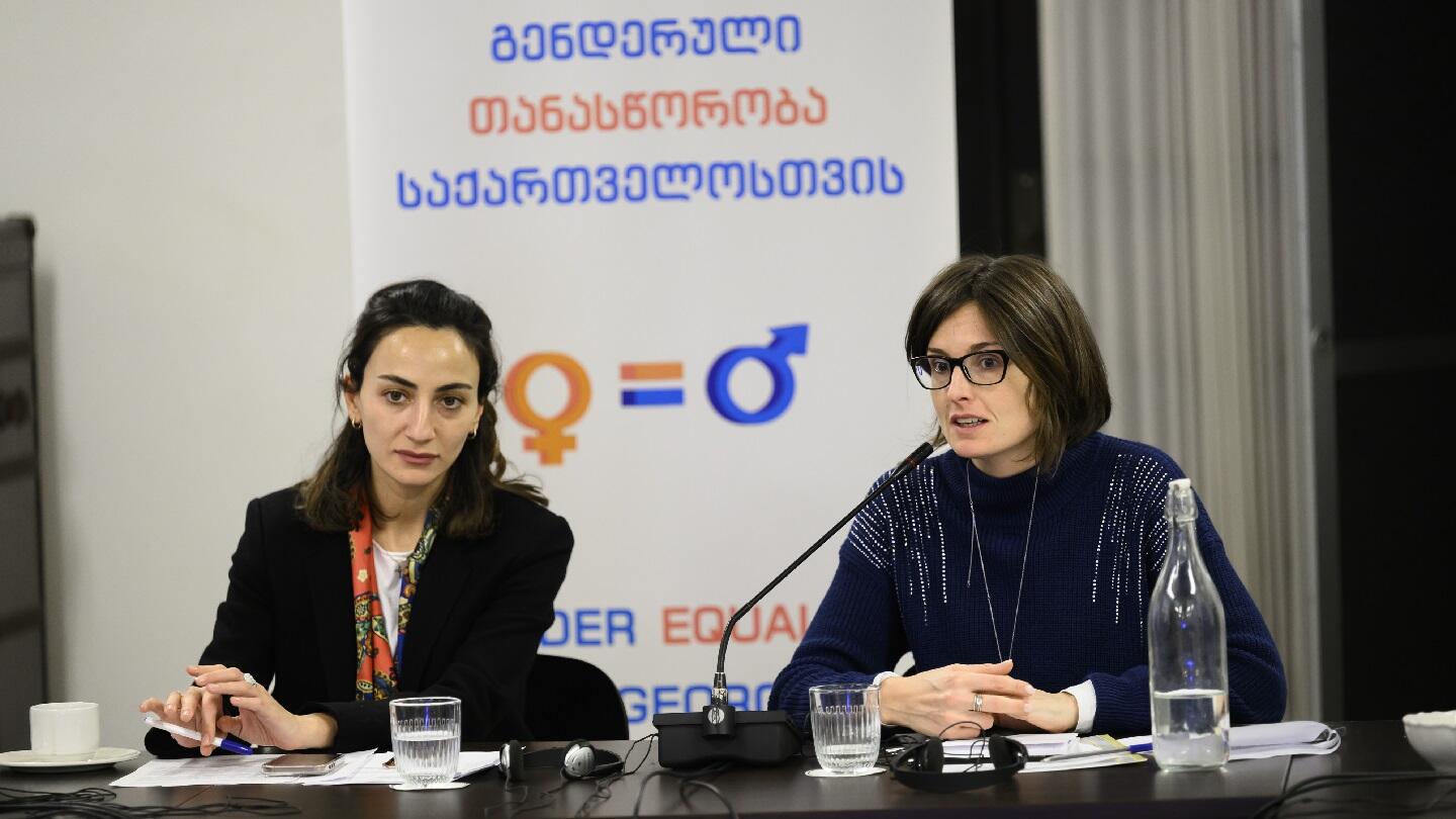 A photo of two women sitting at a table in front of a banner supporting gender equality