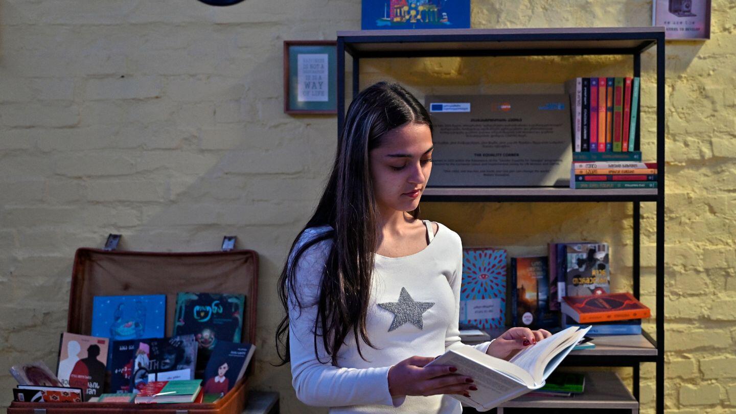 A girl with long hair is standing in front of a bookshelf holding a book