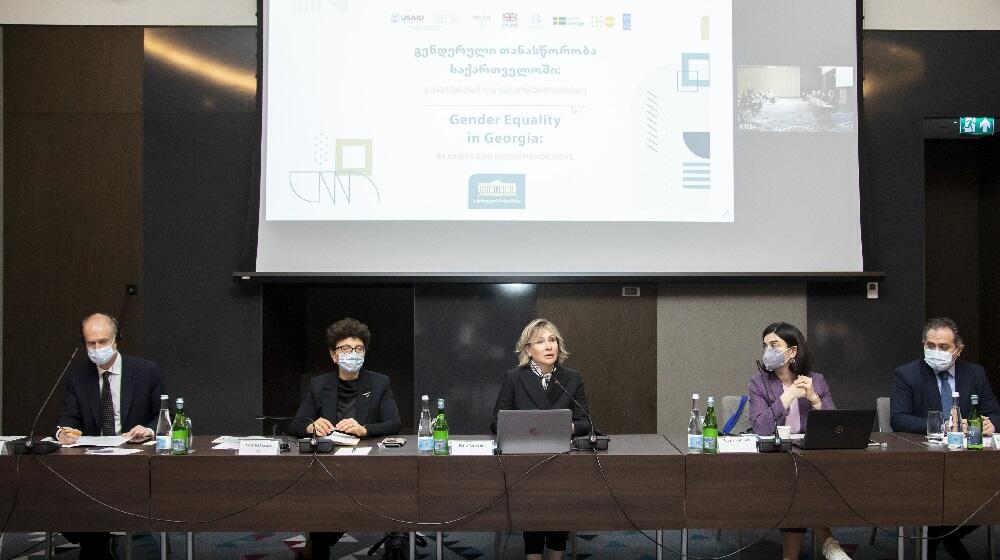 A panel comprised of women and men addresses the audience