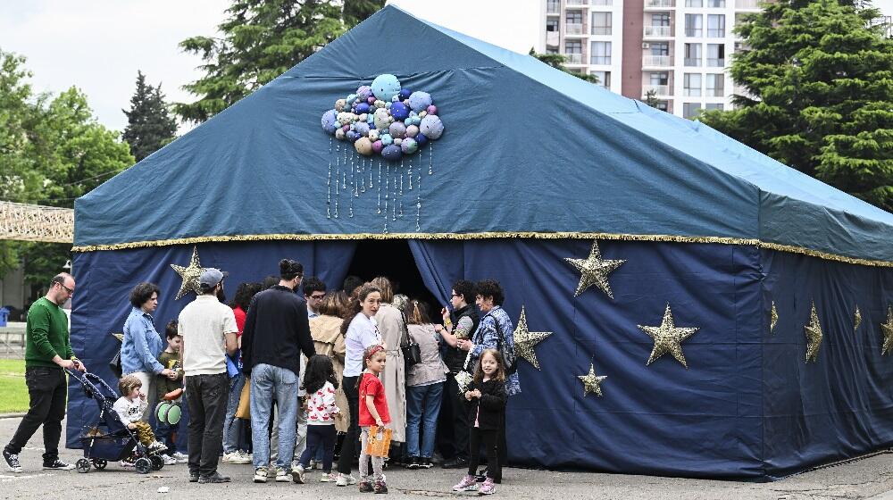 People with kids are standing at a blue tent with yellow start and several balloons