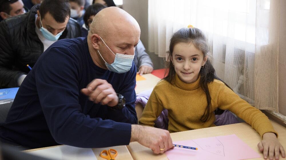 A girl smiling at camera while her dad is helping her paint