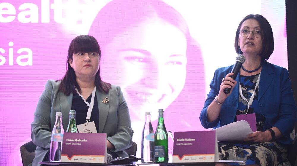 Two women sitting in front of a purple screen, one of them holding a mic