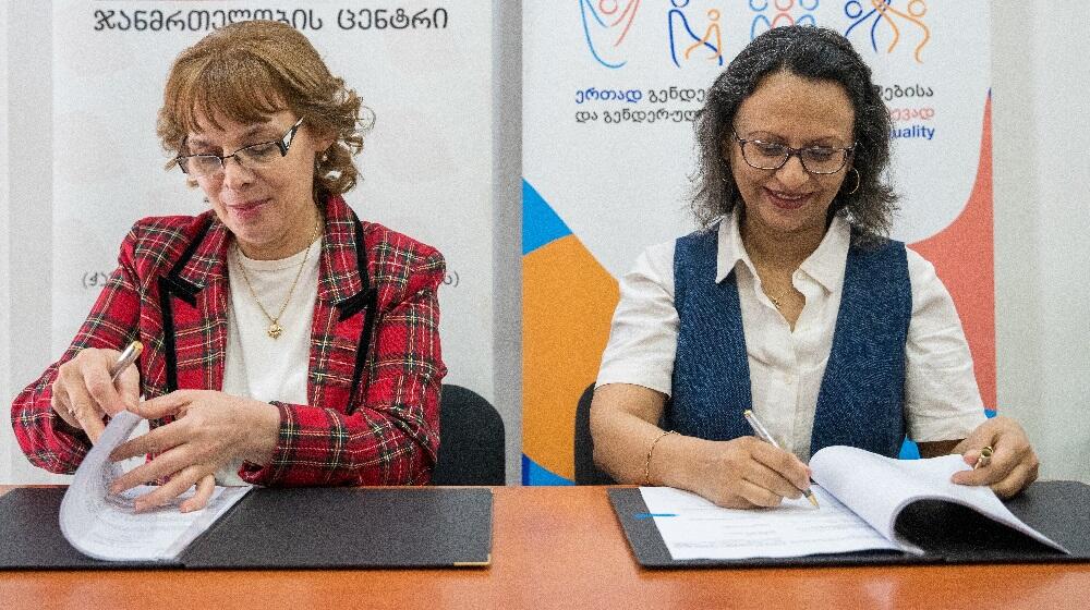 Directors of Hera Clinic and UNFPA Georgia are sitting at a table, signing the memorandums of understanding in front of banners