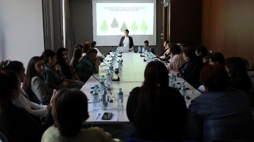 Dozens of women are sitting around a table, listening to a woman, who is standing in front of them, explaining something