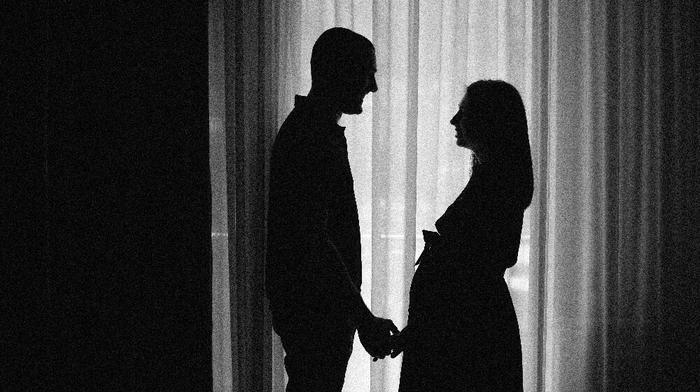 A black and white photo of a couple, expecting a baby standing near the curtains holding hands