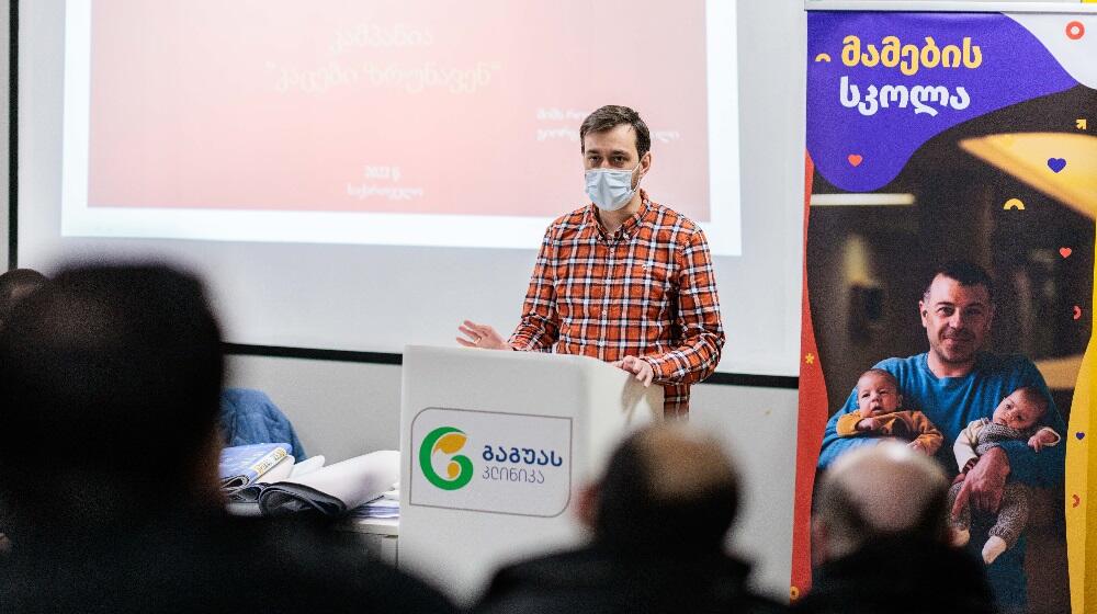 A man wearing a mask is standing in front of a large screen which reads: MenCare campaign. There is a rollup banner of Fathers' Sshool on the left side of the photo